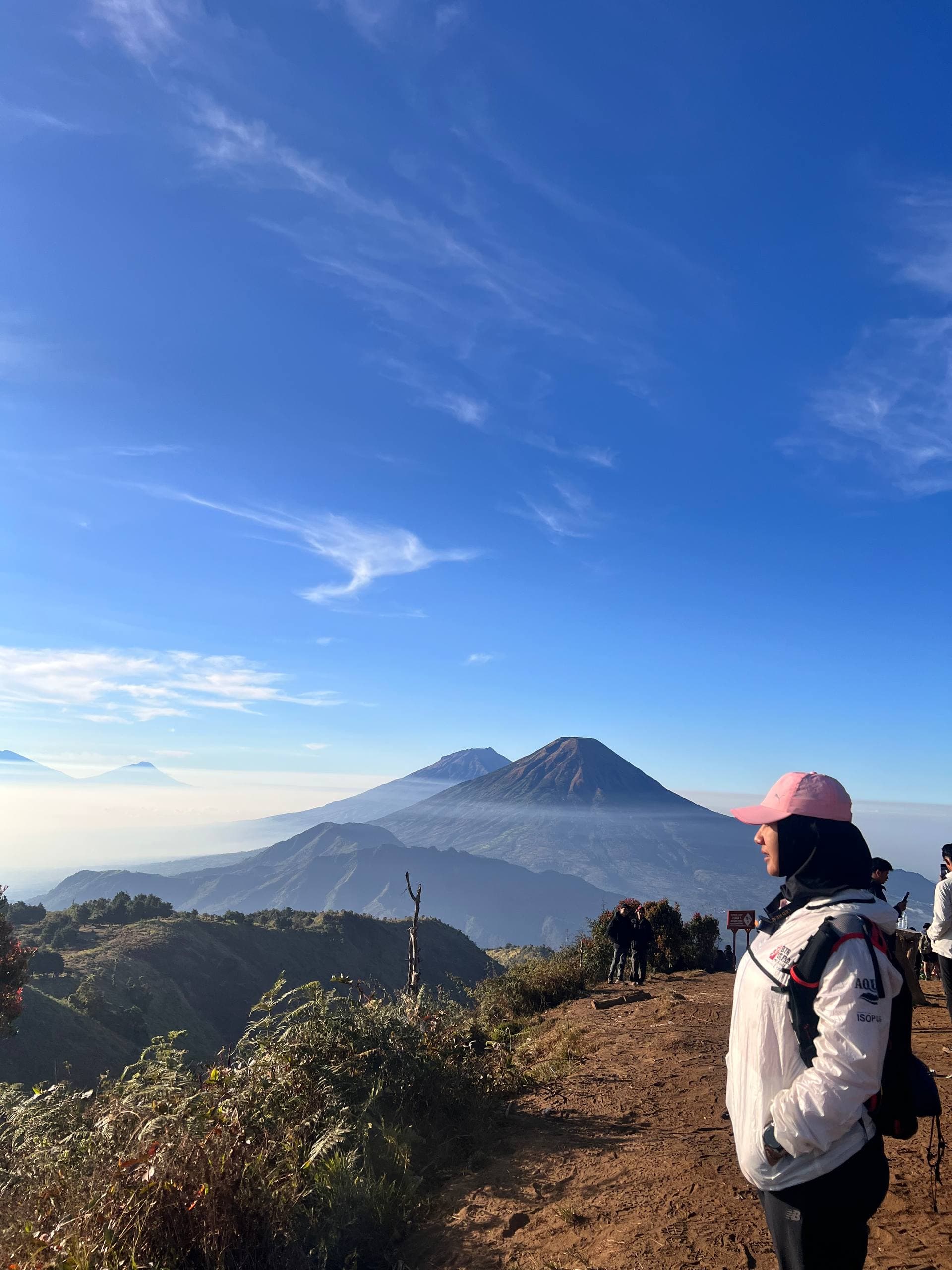 A hiker on the Mount Prau ridge trail at sunrise, with Sindoro and Sumbing rising above a sea of cloud