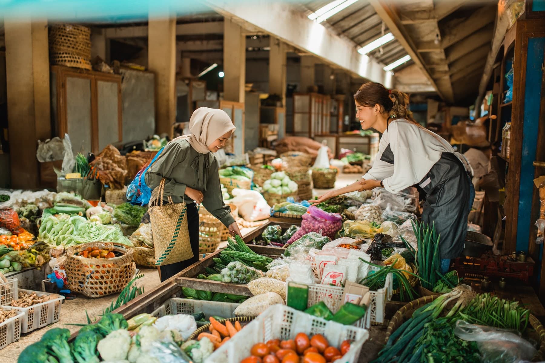 A market seller helping a customer choose vegetables at a stall piled with greens and carrots