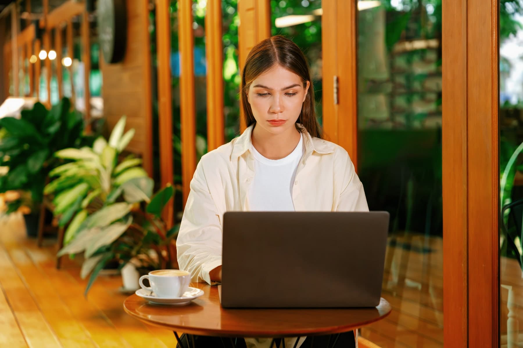 A woman working on a laptop at a cafe table surrounded by plants